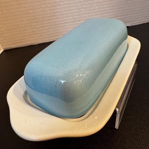 May include: A light blue ceramic butter dish with a matching lid, resting on a cream-colored rectangular base. The dish has a rounded, rectangular shape. The butter dish is a kitchenware item.