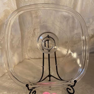 May include: A clear, square glass lid with a circular handle. The lid is displayed on a black metal stand with decorative scrollwork. The background is a light-colored fabric.