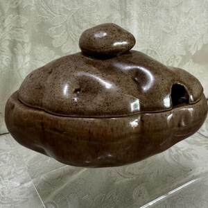 May include: A brown ceramic serving dish shaped like a potato with a lid. The dish has a textured surface and a small hole in the lid.