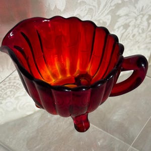 May include: A red glass creamer with a scalloped edge and three legs. The creamer has a handle and is made of ruby red glass.