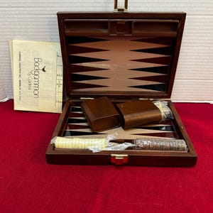May include: A vintage backgammon set in a wooden case, open to reveal the playing board with brown and white triangles. Includes dice cups, dice, and game pieces. The set is on a red surface. The instruction manual reads "Backgammon by Cardinal".
