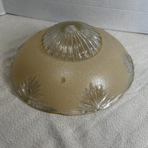 May include: A vintage, frosted glass light fixture shade. The shade is a soft beige color with a textured surface and clear glass accents. The center has a ribbed design, and the edges feature leaf-like patterns.