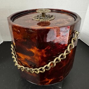 May include: A cylindrical ice bucket with a glossy, marbled brown and orange finish. The lid features a decorative, circular brass-toned knob. A thick, textured brass-toned metal chain is attached to the sides, serving as a functional handle.
