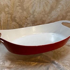 May include: A red and white oval baking dish with handles. The exterior is a glossy red, while the interior is white. The dish has a curved shape and is designed for oven use. This kitchenware item is suitable for baking and serving.