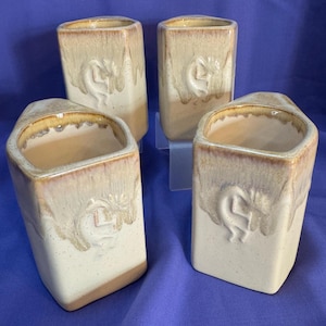 May include: Set of four ceramic mugs with a beige and brown glaze. Each mug features a raised design of a Native American dancer.