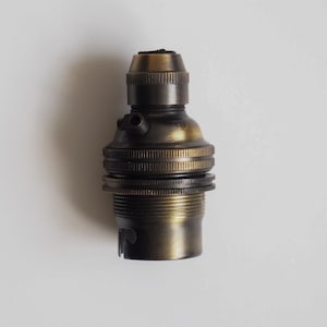 May include: A brass lamp holder with a vintage aesthetic. The lamp holder features a ridged design and a cylindrical shape. It has a threaded base and a narrower top section. The brass finish has a slightly aged appearance.