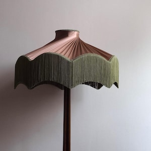 May include: A vintage-style floor lamp with a pink lampshade and olive green fringe. The lampshade has a scalloped edge and is supported by a dark brown wooden pole. The lamp is a decorative lighting fixture.