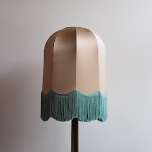 May include: A vintage-style lampshade with a beige, dome-shaped top and a scalloped edge. The edge is trimmed with a fringe of teal-colored tassels. The lampshade is mounted on a dark brown wooden pole.