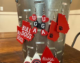 May include: A collection of red and white painted wooden earrings with various designs. The earrings are displayed on a metal bucket. Some of the designs include the letters "UGA", "Bulldogs", and "Dawgs".