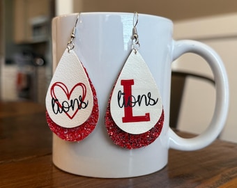 Pode incluir: Um par de brincos em forma de lágrima vermelha e branca com glitter. Um brinco tem um coração vermelho com a palavra "lions" escrita em preto. O outro brinco tem uma letra "L" vermelha com a palavra "lions" escrita em preto.