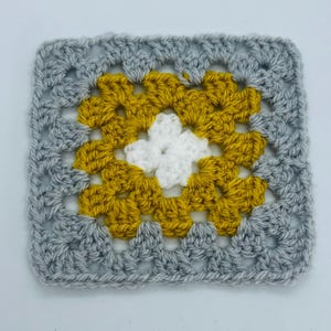 May include: A crocheted square with a white center surrounded by a yellow border and a gray outer border.
