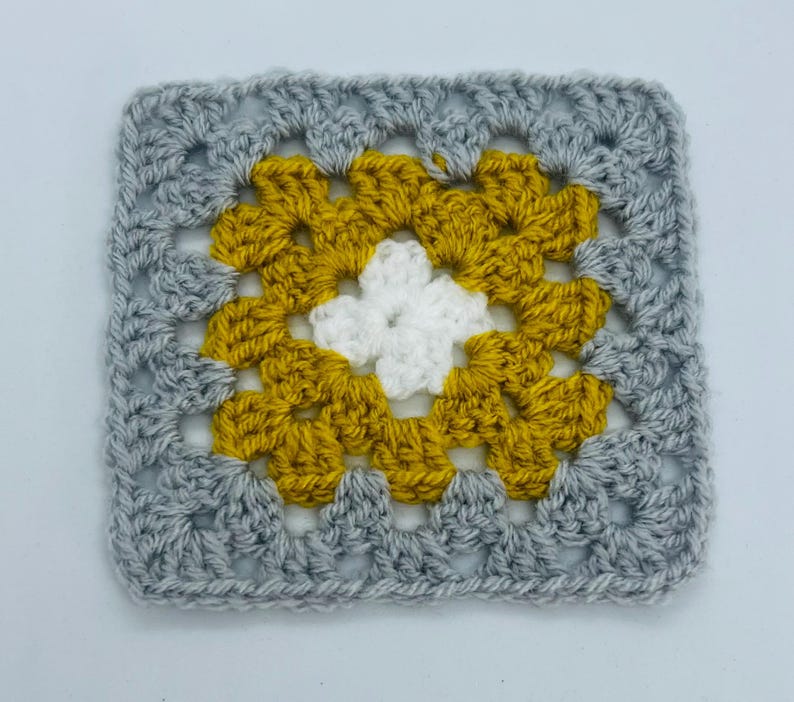 May include: A crocheted square with a white center surrounded by a yellow border and a gray outer border.