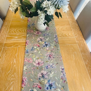 May include: A grey floral table runner with a watercolour design featuring pink, blue and green flowers. The runner is on a wooden table with a vase of white and blue flowers.