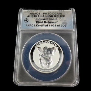 May include: A silver coin featuring a koala bear clutching a tree branch, with the words "AUSTRALIAN KOALA" around the coin's edge. The coin is encased in a clear plastic holder with text that reads "ANACS - PR70 DCAM AUSTRALIA HIGH RELIEF Incused Koala First Release".