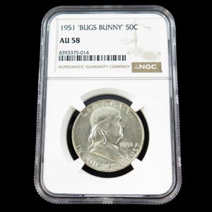 May include: A silver 1951 50-cent coin, graded AU 58, encased in a clear protective holder. The coin features a profile of a man and the text "IN GOD WE TRUST". The holder has the text "1951 'BUGS BUNNY' 50C" and "NUMISMATIC GUARANTY COMPANY".