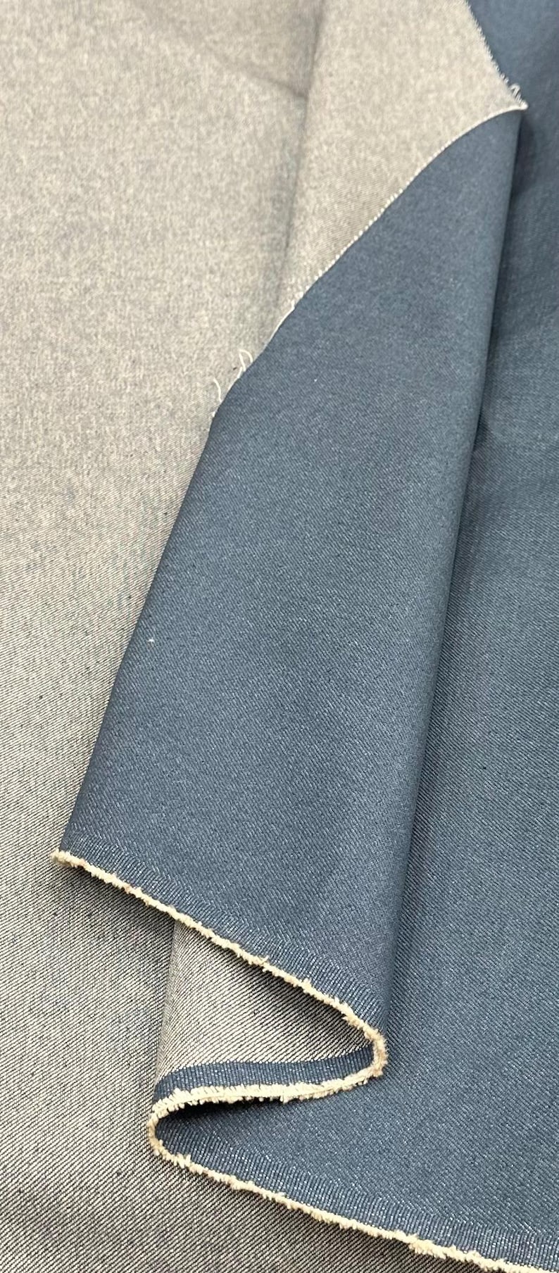 May include: A piece of folded fabric featuring a dual-sided design with a dark blue, smooth-textured side and a lighter, heathered gray side. The material shows a visible weave pattern and raw, frayed edges along the folded section of the textile.