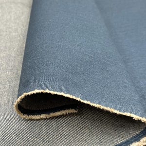 May include: A roll of dark blue denim fabric featuring a distinct twill weave pattern. The material has a folded edge that reveals a lighter, beige-colored underside with a raw, frayed texture along the selvage of the heavy-duty textile.