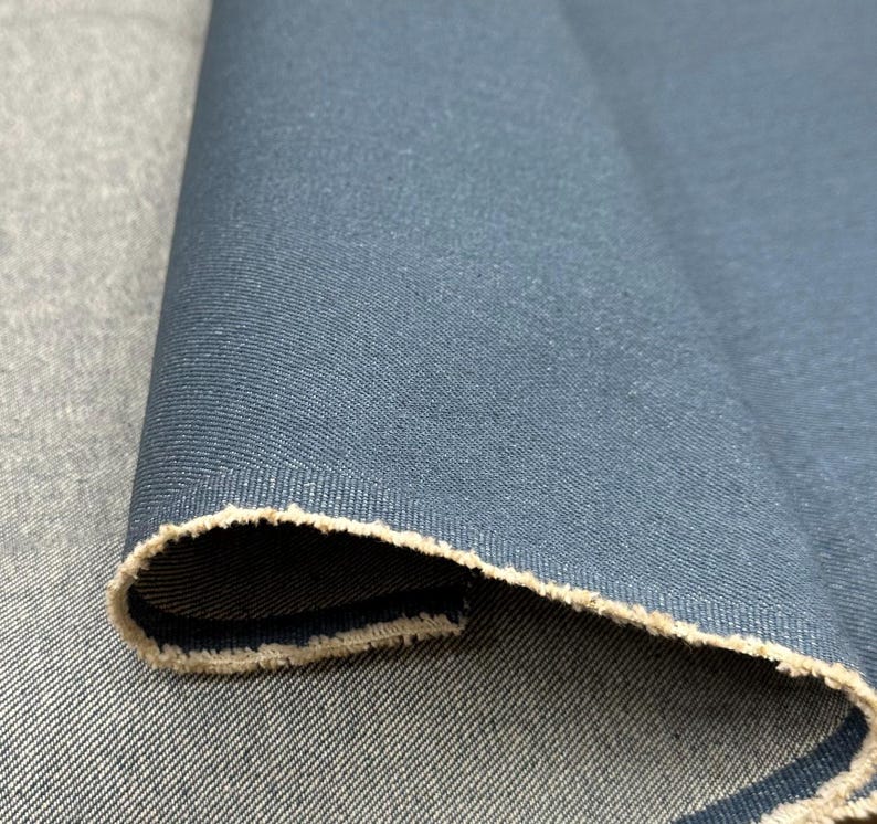 May include: A roll of dark blue denim fabric featuring a distinct twill weave pattern. The material has a folded edge that reveals a lighter, beige-colored underside with a raw, frayed texture along the selvage of the heavy-duty textile.