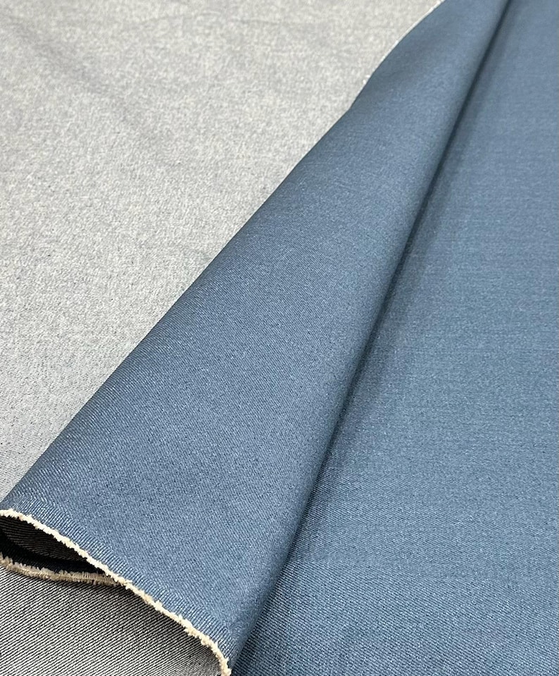 May include: A folded piece of fabric featuring a deep blue side with a smooth, matte texture and a contrasting light gray, heathered underside. The material displays a visible woven grain and a raw, frayed edge along the diagonal fold line.