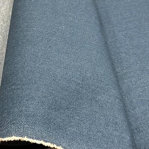 May include: A length of dark blue denim fabric featuring a visible twill weave pattern. The material is folded to show the textured surface and the raw, frayed edge along the bottom, revealing a lighter beige color on the underside of the fabric.