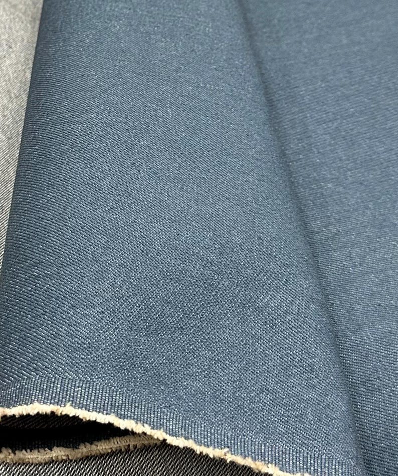 May include: A length of dark blue denim fabric featuring a visible twill weave pattern. The material is folded to show the textured surface and the raw, frayed edge along the bottom, revealing a lighter beige color on the underside of the fabric.