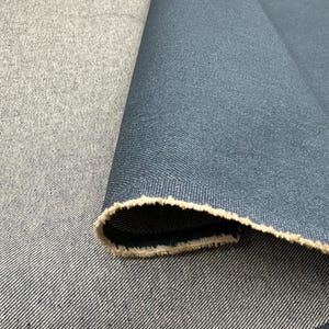 May include: A roll of dark blue denim fabric with a visible twill weave pattern. The fabric is folded over to reveal a lighter, uncolored beige interior edge with a frayed, raw texture along the selvage, showcasing the dense textile construction.