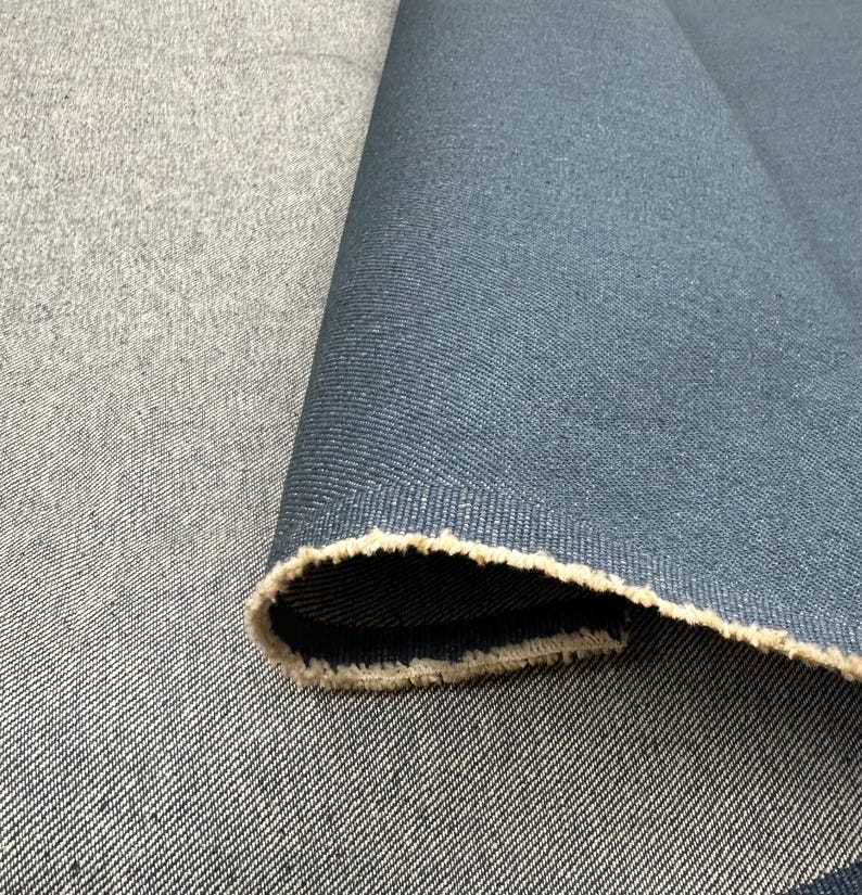 May include: A roll of dark blue denim fabric with a visible twill weave pattern. The fabric is folded over to reveal a lighter, uncolored beige interior edge with a frayed, raw texture along the selvage, showcasing the dense textile construction.