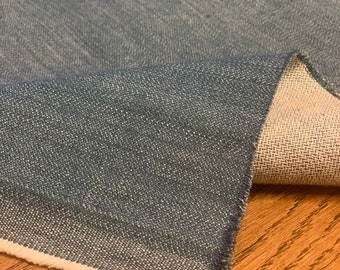 May include: A close-up of a piece of blue denim fabric with a white selvedge edge. The fabric has a textured weave and a slightly faded appearance.