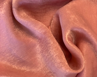 May include: Close-up of a flowing, blush-colored fabric with a subtle sheen. The material is soft and appears to be draped, with gentle folds and highlights. The texture suggests a luxurious textile, possibly silk or satin.