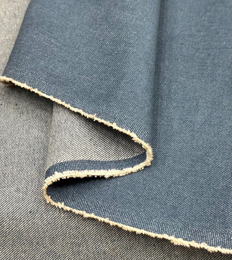 May include: A piece of dark blue denim fabric with a visible twill weave pattern. The material is folded over to reveal a lighter, off-white underside with a raw, frayed edge along the fold, showcasing the contrast between the two sides.
