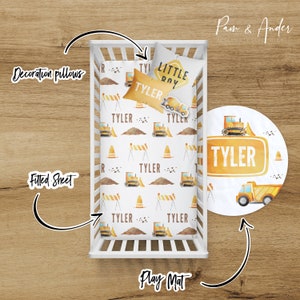 May include: A white crib with a fitted sheet and a decorative pillow with a construction theme. The sheet has a pattern of yellow construction vehicles, cones, and barriers. The pillow has the words "Little Boy" and "Tyler" on it. There is also a round play mat with a yellow construction truck and the name "Tyler" on it.