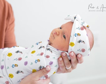 Op de afbeelding: Een witte swaddle met een herhalend patroon van Alice in Wonderland-personages en gele bloemen. Een baby is gewikkeld in de swaddle en draagt ​​een bijpassend hoofdband met hetzelfde patroon. Het hoofdband heeft een strik bovenop.