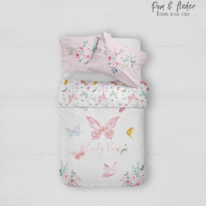 Puede incluir: Un juego de cama blanco con un estampado floral rosa y verde y mariposas. El juego de cama incluye una funda nórdica y dos fundas de almohada. La funda nórdica tiene el nombre "Emily Rose" escrito en ella en letras cursivas rosas.
