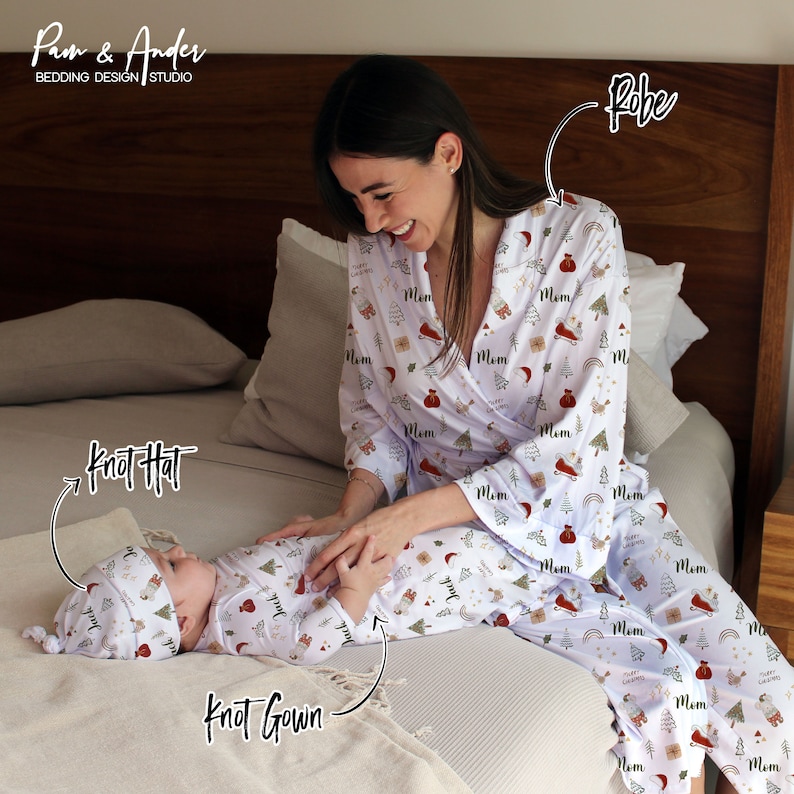 May include: A white robe with a Christmas print featuring red and green accents. The robe has the word "Mom" printed on it. A baby is wearing a white knotted gown with a matching Christmas print and a knotted hat. The baby is lying on a bed with a white blanket.