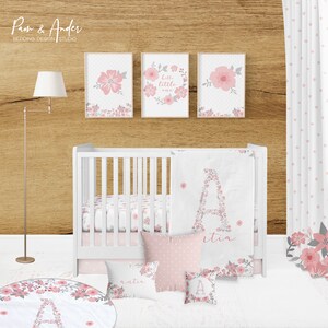 May include: A white crib with pink and gray floral bedding. The crib has a white blanket with a floral letter A on it. There are two pink pillows with floral designs and a pink pillow with white polka dots. There is a pink and gray floral rug on the floor. The wall behind the crib is wood and has three framed prints with pink and gray floral designs. The text on one of the prints says "hello little one".