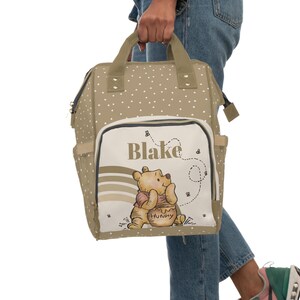 May include: A beige diaper bag with white polka dots and a Winnie the Pooh design. The bag has a large front pocket with the name "Blake" and a picture of Winnie the Pooh holding a jar of honey.