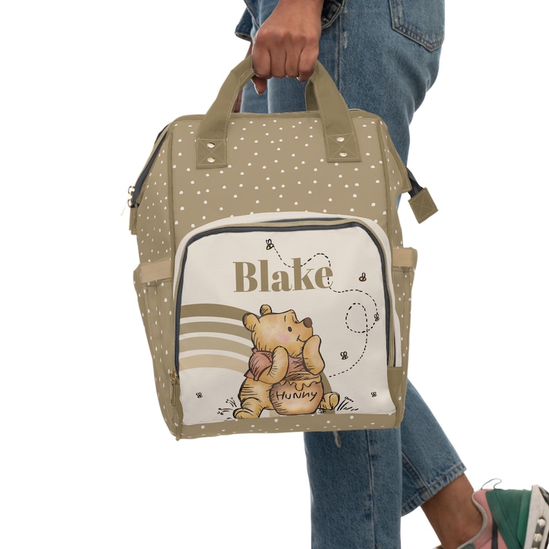 May include: A beige diaper bag with white polka dots and a Winnie the Pooh design. The bag has a large front pocket with the name "Blake" and a picture of Winnie the Pooh holding a jar of honey.