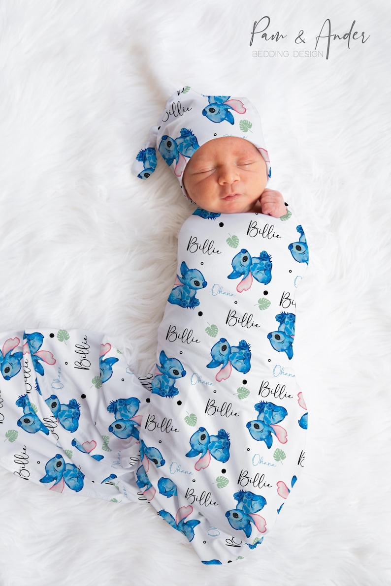 May include: A white swaddle blanket with a blue and pink Stitch pattern. The blanket has the name "Billie" and the word "Ohana" printed on it. A baby is wearing a matching blue and pink Stitch hat.