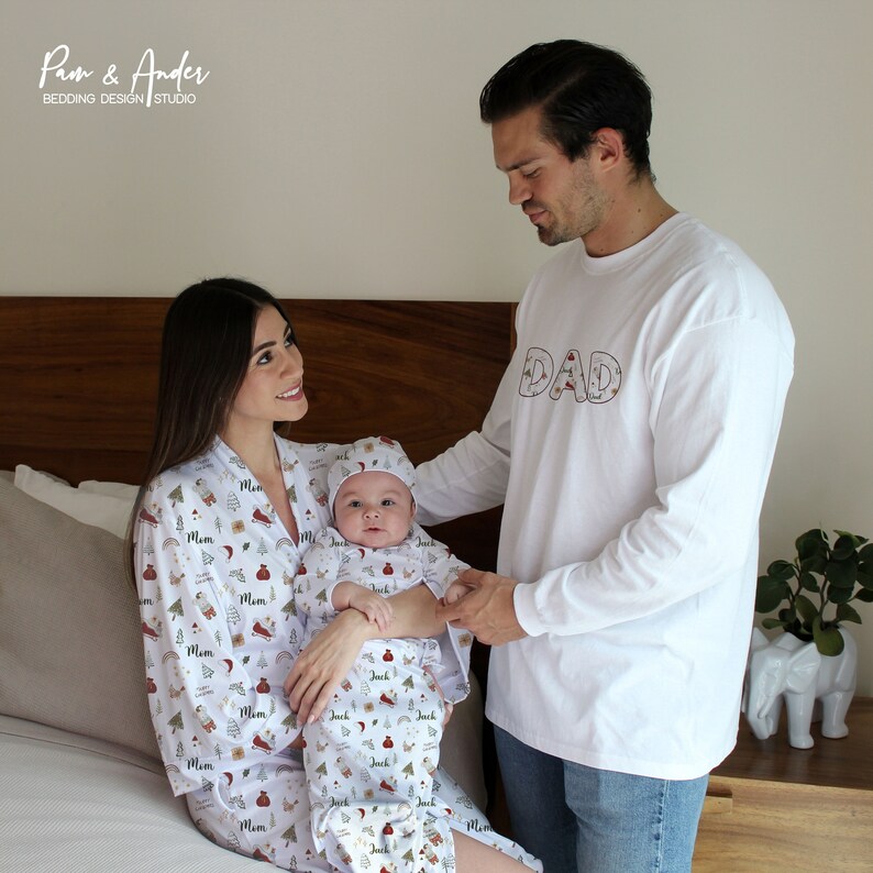 May include: A family wearing matching white pajamas with a Christmas-themed print. The pajamas feature a red and green pattern with illustrations of Christmas trees, presents, and ornaments. The father's pajamas have the word "DAD" written on the front. The mother's pajamas have the word "MOM" written on the front. The baby's pajamas have the word "JACK" written on the front.