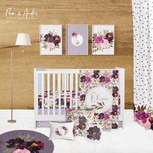 May include: A white cot with a floral patterned cot bedding set. The bedding includes a fitted sheet, a cot skirt, a blanket, and two pillows. The cot is decorated with a floral wreath and the name "Olivia" in a circle. The cot is in a nursery with a wooden wall and a white curtain with purple polka dots.