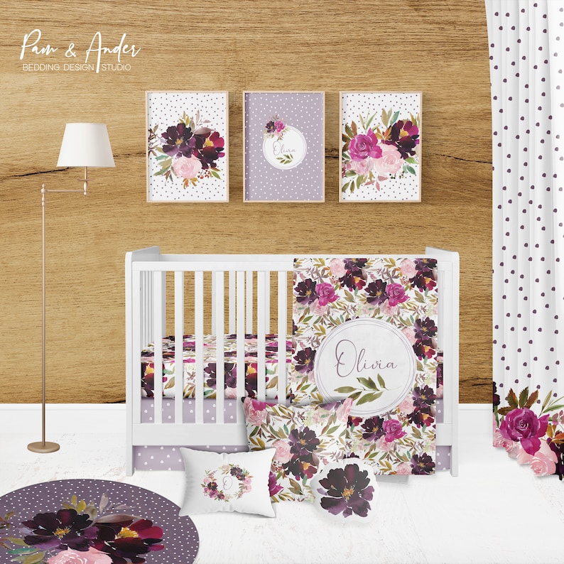 May include: A white cot with a floral patterned cot bedding set. The bedding includes a fitted sheet, a cot skirt, a blanket, and two pillows. The cot is decorated with a floral wreath and the name "Olivia" in a circle. The cot is in a nursery with a wooden wall and a white curtain with purple polka dots.
