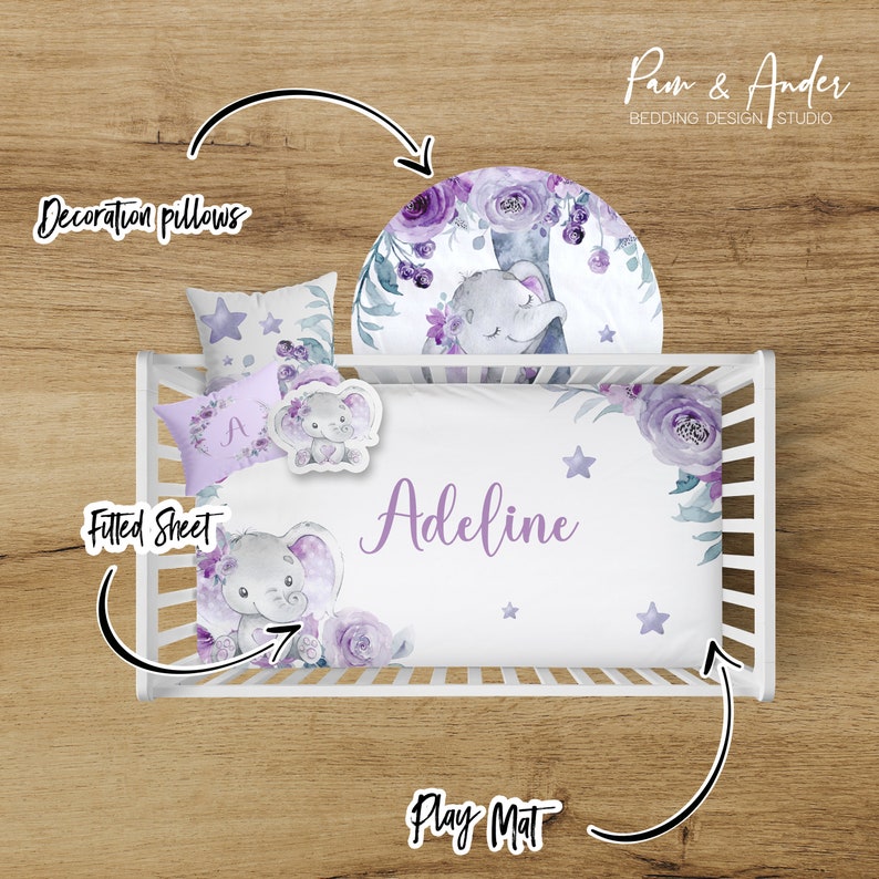 May include: A white cot bedding set with a purple floral and elephant design. The set includes a fitted sheet, a decorative pillow, and a play mat. The fitted sheet has the name "Adeline" embroidered on it.