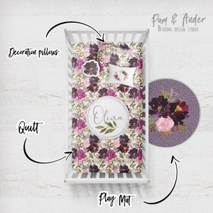 May include: A white cot with a floral patterned quilt and a matching play mat. The quilt and play mat feature a burgundy, pink, and green floral design. The play mat is round and has a purple background with white polka dots. The cot also includes a decorative pillow with a floral wreath design. The text "Olivia" is printed on the quilt in a circle. The text "Pam & Ander Bedding Design Studio" is printed in the top right corner of the image.