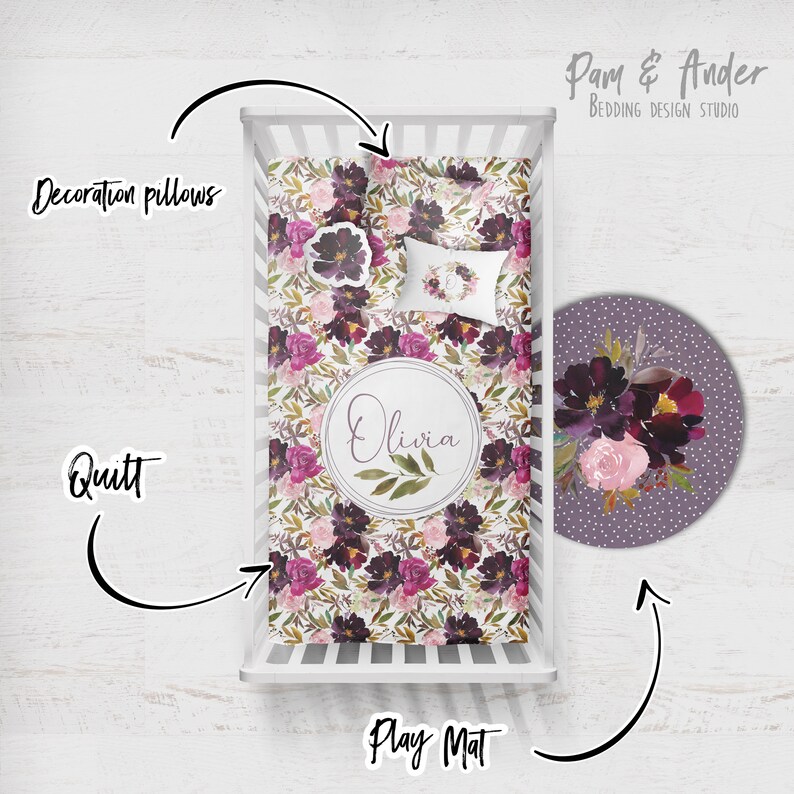 May include: A white cot with a floral patterned quilt and a matching play mat. The quilt and play mat feature a burgundy, pink, and green floral design. The play mat is round and has a purple background with white polka dots. The cot also includes a decorative pillow with a floral wreath design. The text "Olivia" is printed on the quilt in a circle. The text "Pam & Ander Bedding Design Studio" is printed in the top right corner of the image.