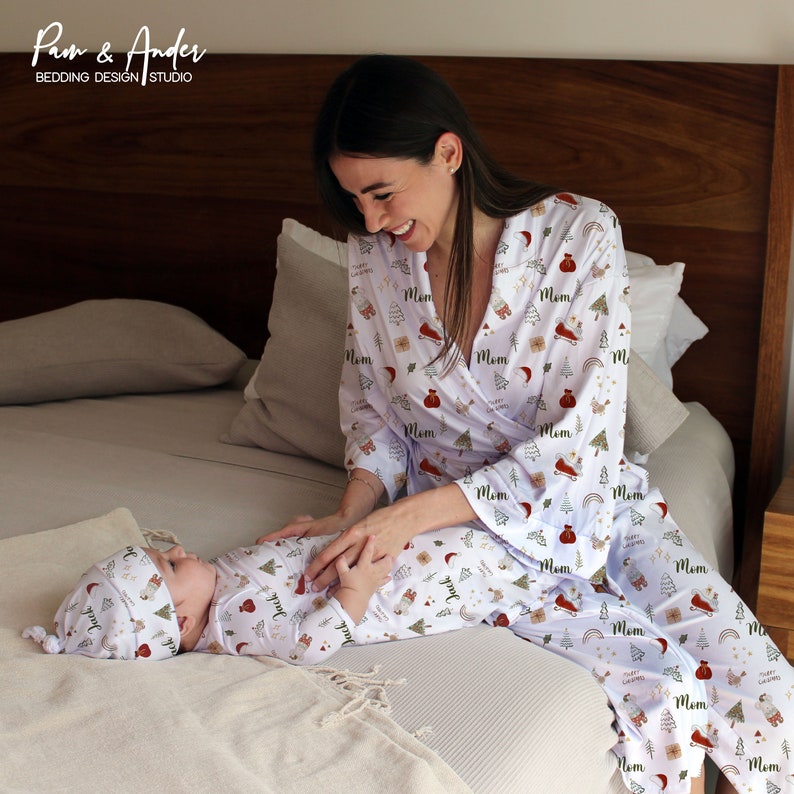 May include: A mother and child wearing matching white pajamas with a Christmas-themed print. The pajamas feature a white background with red and green illustrations of Christmas trees, presents, and the word "Mom". The child is wearing a matching hat.