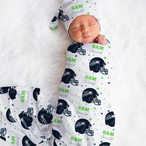 May include: A newborn baby wrapped in a white swaddle and matching hat, both patterned with navy blue football helmets, green stars, and the name "SAM Darnold". The bedding design is by Pam & Ander.