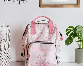 May include: A pink and white floral patterned diaper bag backpack with the name "Marianne" printed on the front. The bag has a zippered front pocket and adjustable straps.