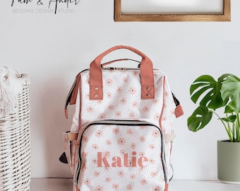May include: A pink and white floral patterned diaper bag with the name "Katie" in pink lettering. The bag has a top handle and adjustable straps.