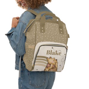 May include: A beige and brown Winnie the Pooh diaper bag backpack with white polka dots. The bag has a large pocket on the front with a Winnie the Pooh illustration and the name "Blake" printed on it.