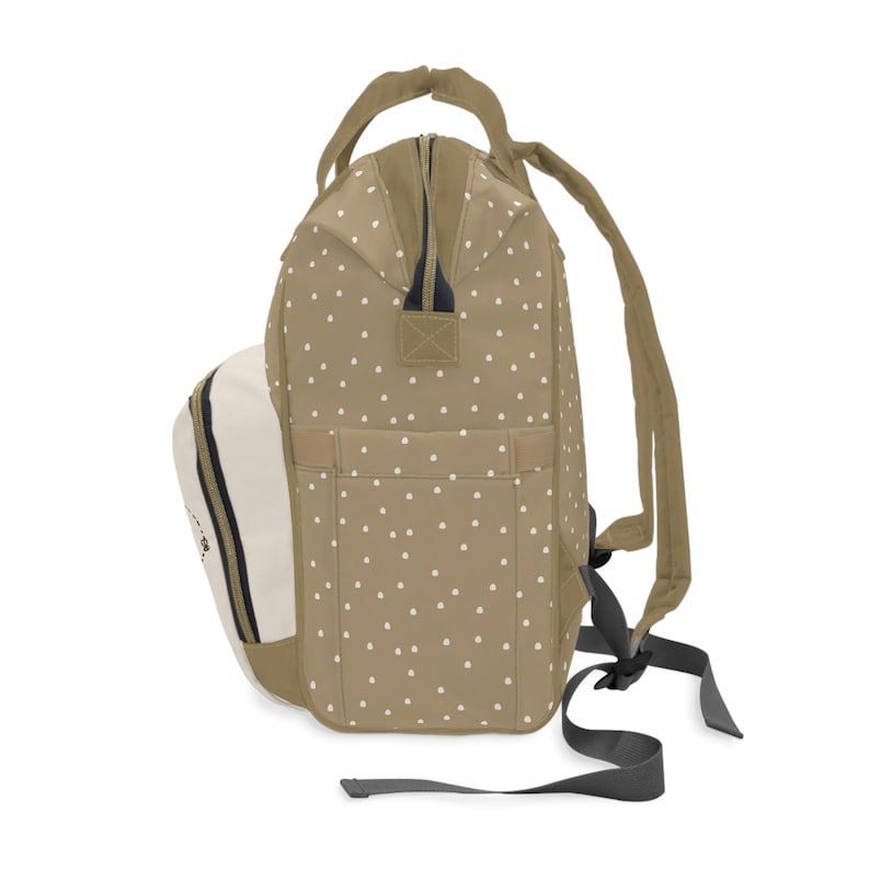 May include: A beige and white polka dot backpack with a white and black zipper. The backpack has a large main compartment and a smaller front pocket. The straps are adjustable and have a padded shoulder area.